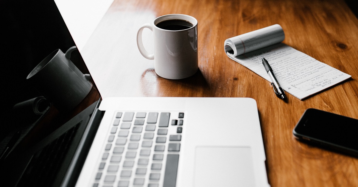 wooden desk with a laptop, coffee cup and notpad with pen