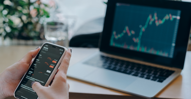 Someone holding their phone at a desk with a laptop showing a stock ticker