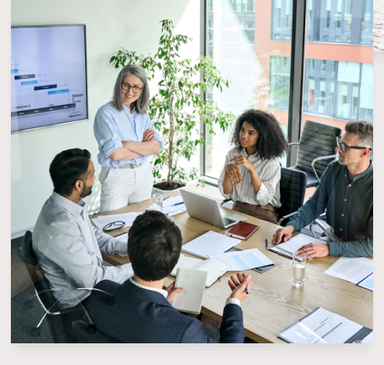 A business meeting with several people sitting around a table with one standing