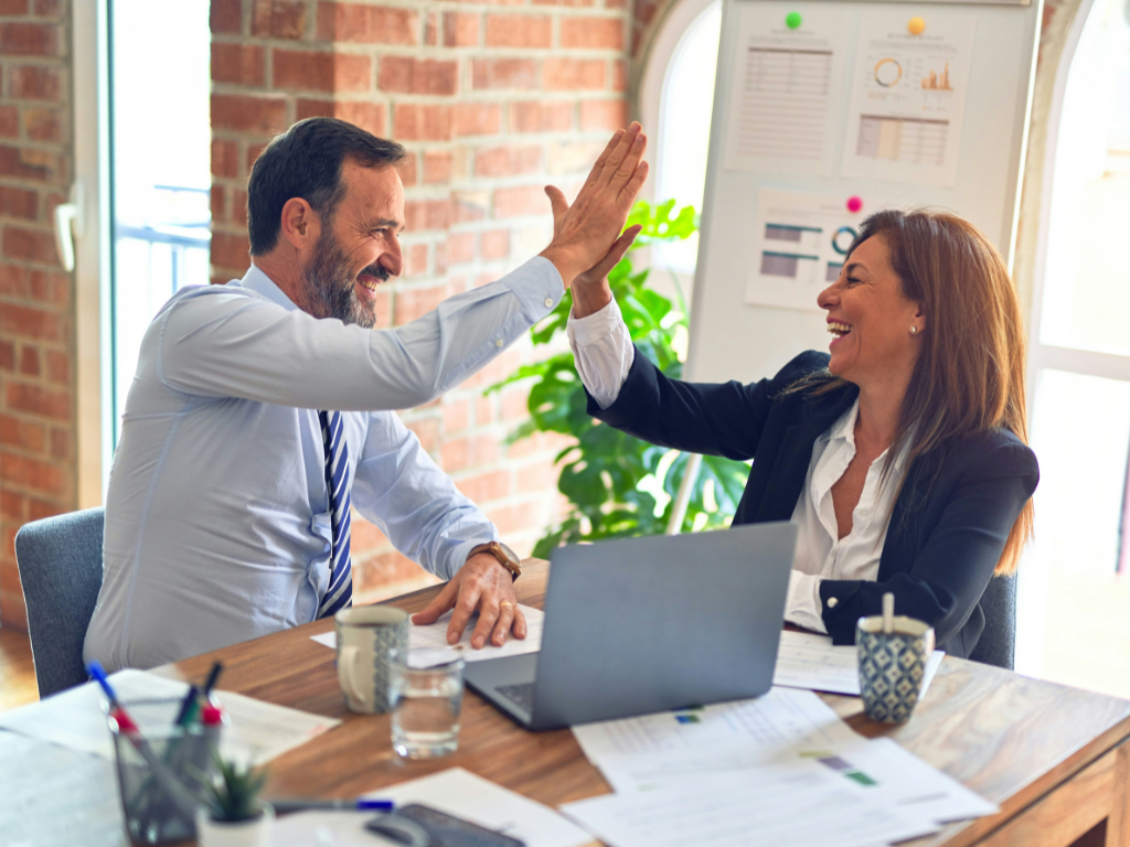 A man and woman high fiving at a desk with a laptop, coffee mugs and papers