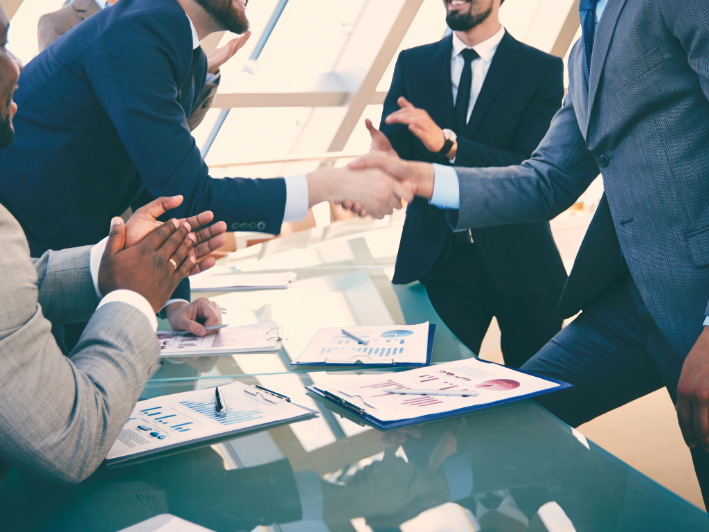 Two men shaking hands over a desk with papers on them and others applauding