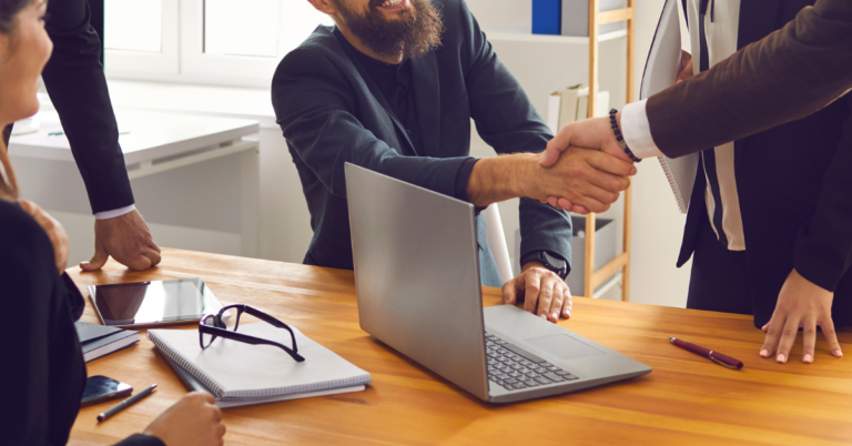 Shaking hands with other people around a table with a laptop and notepad on it